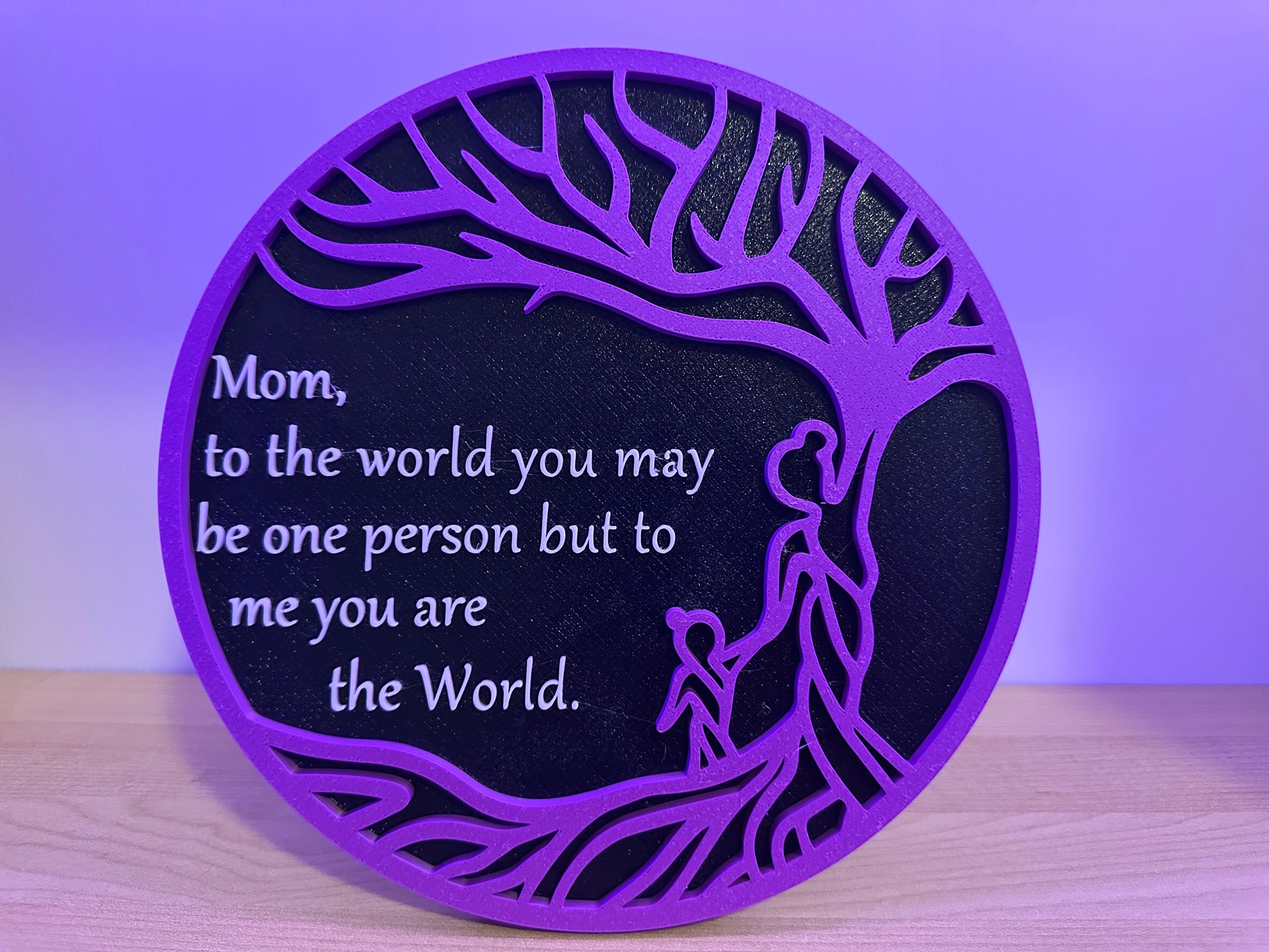 Decorative plaque with purple border and black center featuring a tree and Mom you are the world, on a wooden surface with a purple background.