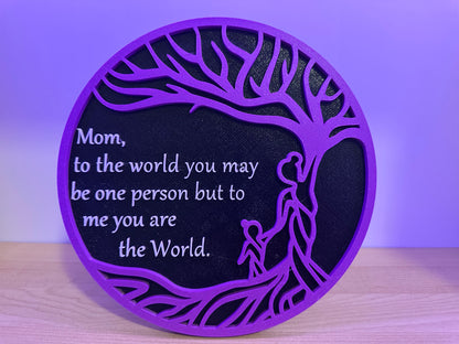 Decorative plaque with purple border and black center featuring a tree and Mom you are the world, on a wooden surface with a purple background.